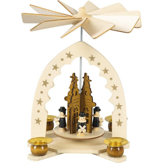 Naturfarbene Weihnachtspyramide mit Kurrende. Betrieben wird die Pyramide mit 4 Pyramidenkerzen. Der Durchmesser der Kerzen beträgt 1,4 cm. Auf der Pyramide befindet sich eine Kirche. Vor dieser Kirche stehen die Kurrendesänger mit Ihren Gasangsbüchern in der Hand. Die Tüllen sind farbig abgesetzt und auf dem Bogen befinden sich Sterne.